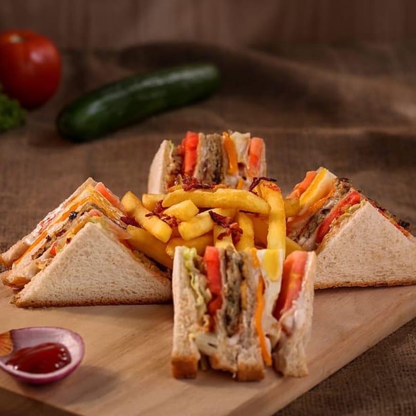Club sandwiches with fries and ketchup on a wooden board, with vegetables in the background.