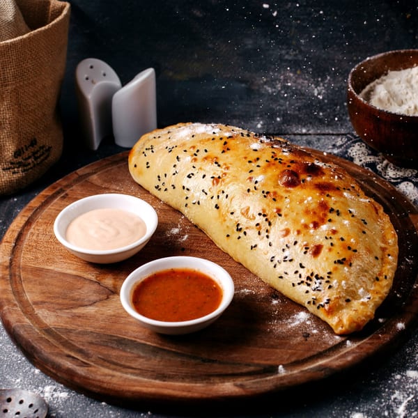 Golden-brown calzone with black sesame seeds, served with two dipping sauces on a wooden board.