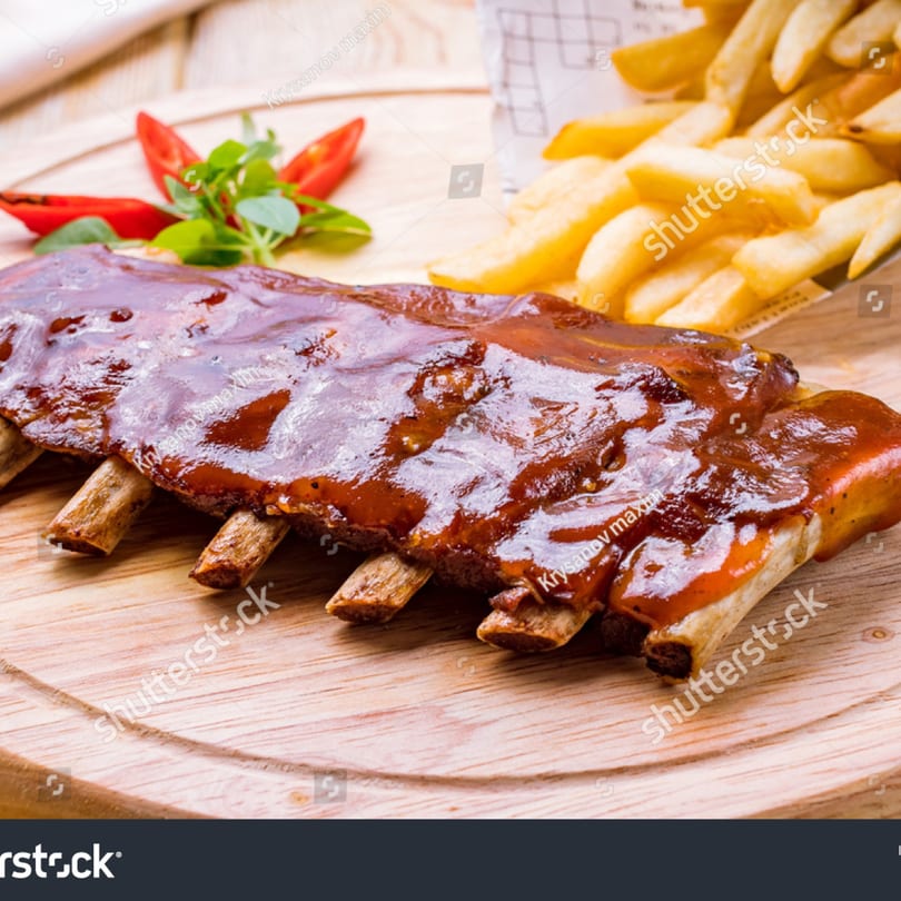 Rack of BBQ ribs with french fries, garnished with red peppers and greens on a wooden board.